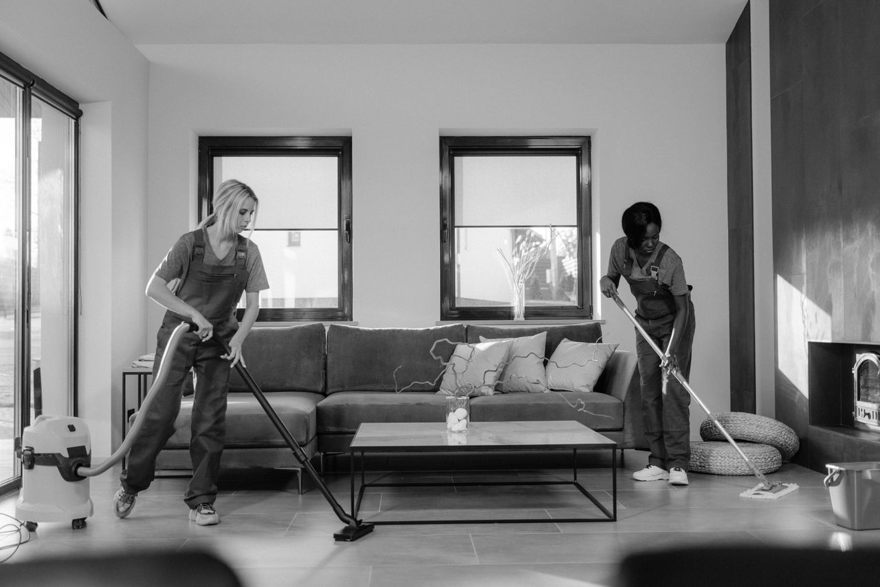 why-choose-us Deux femmes en tenue de travail nettoient un salon moderne à l'aide d'un aspirateur et d'une serpillière.